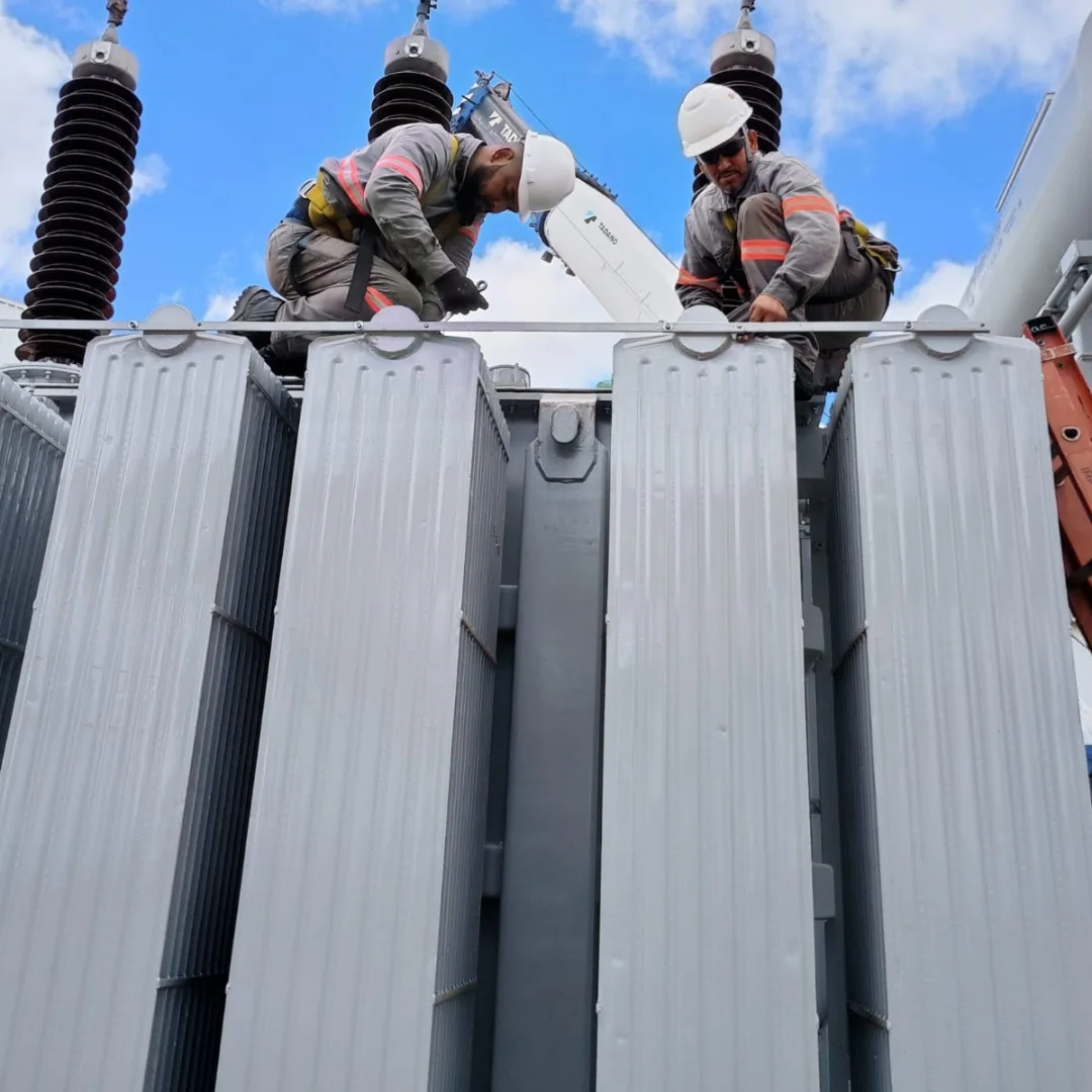 Dois técnicos da Geraenergy com macacão refletivo, capacete e cinto de segurança trabalhando sobre os radiadores de um transformador de potência, com as buchas de alta tensão em primeiro plano